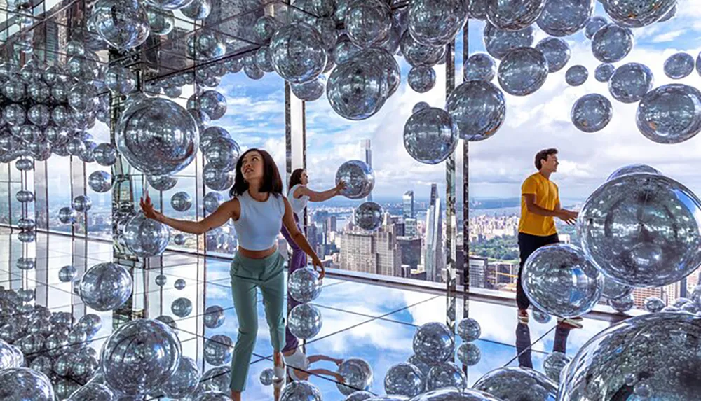 Visitors explore an artistic installation featuring numerous suspended reflective spheres with a panoramic city view in the background