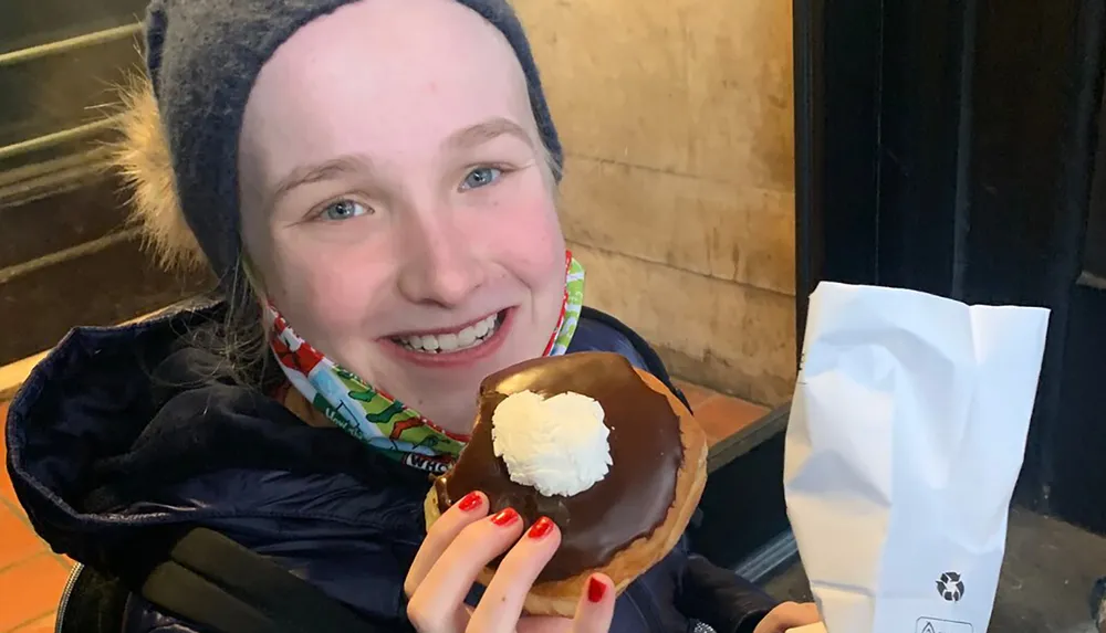 A smiling person is holding a large chocolate-covered donut with a dollop of cream in the center