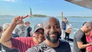 Two people are smiling for a selfie with the Statue of Liberty in the background, while one person playfully pretends to touch the statue with their finger.