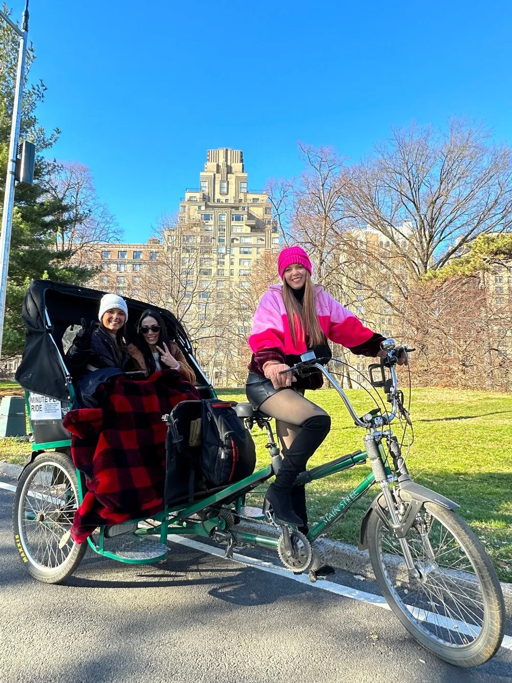A rickshaw cyclist wearing a bright pink beanie is posing for a photo while two passengers are seated in the carriage enjoying a sunny day outdoors with a backdrop of bare trees and a tall building