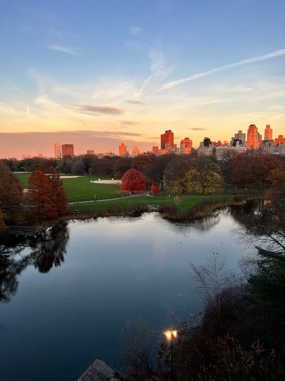 The image captures a serene sunset with a captivating interplay of light and shadow over a park with a pond surrounded by autumn-colored trees and urban high-rises in the background
