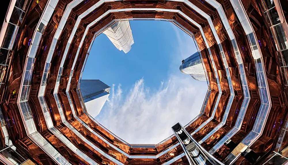 The image shows an upward view from the center of a large intricate hive-like structure with mirrored surfaces reflecting the surrounding skyscrapers and blue sky