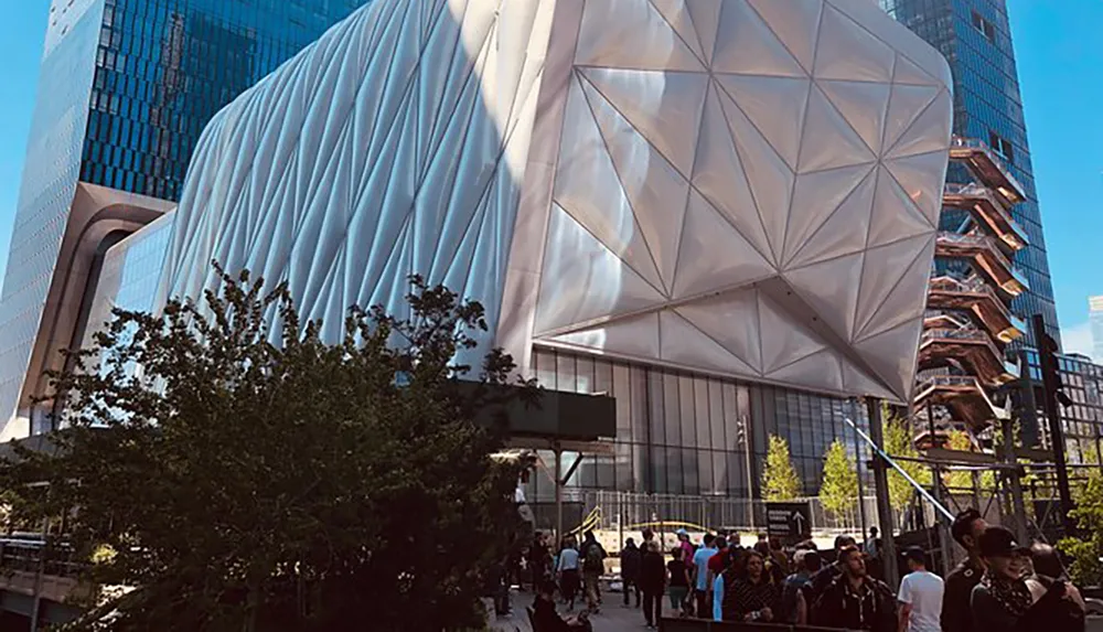 The image shows a modern urban landscape with people walking near a distinctive futuristic structure with an irregular faceted facade flanked by a high-rise building and the Vessel structure at the Hudson Yards in New York City