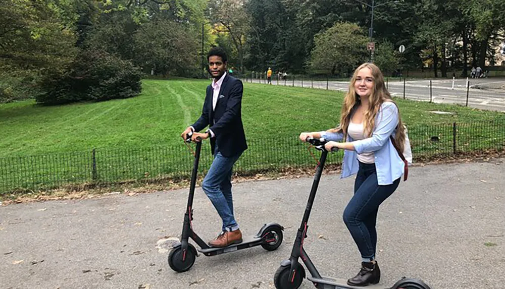 A man and a woman are enjoying a ride on electric scooters along a paved path in a park with green lawns and trees in the background