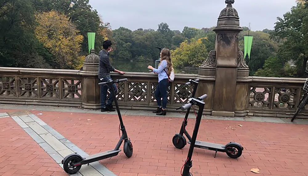 Two people are conversing by a balustrade overlooking a park with trees in autumn colors with two electric scooters parked in the foreground