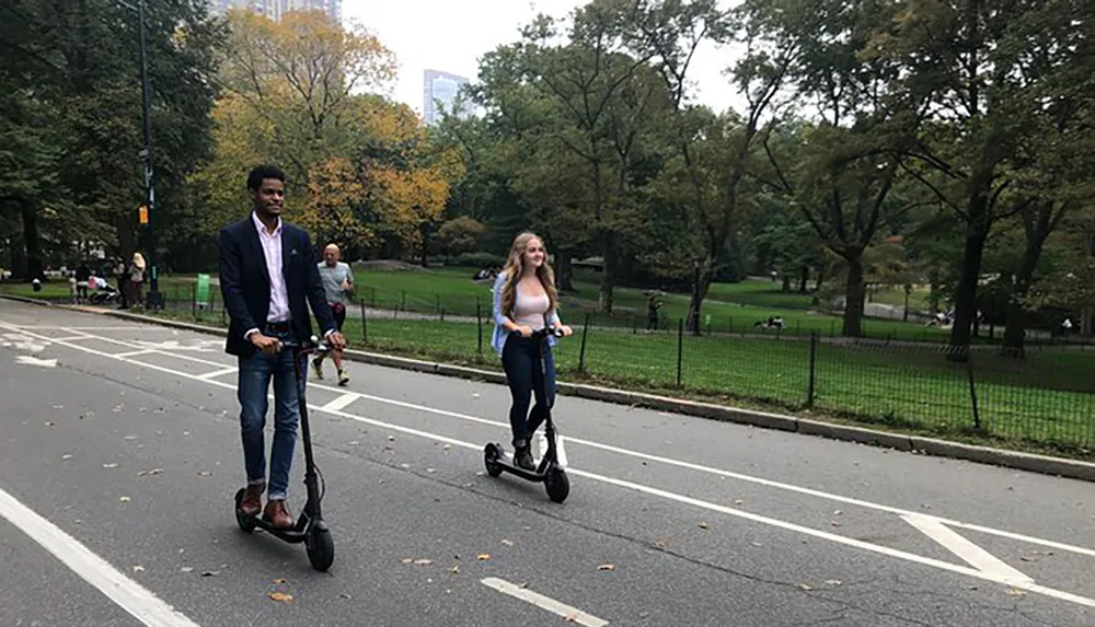 Two people are riding electric scooters along a park path surrounded by trees and grass