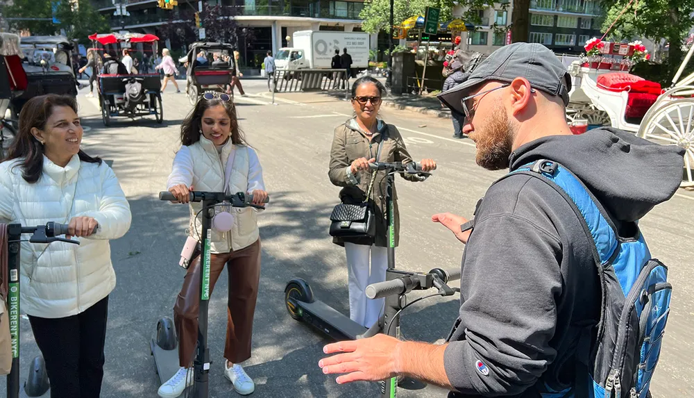 A man appears to be giving instructions or guidance to three women who are ready to ride electric scooters on a sunny day with horse-drawn carriages visible in the background