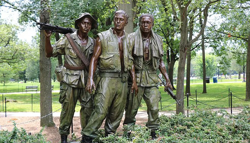 The image shows a bronze statue of three soldiers likely representing a war memorial set against a backdrop of trees in a park-like setting