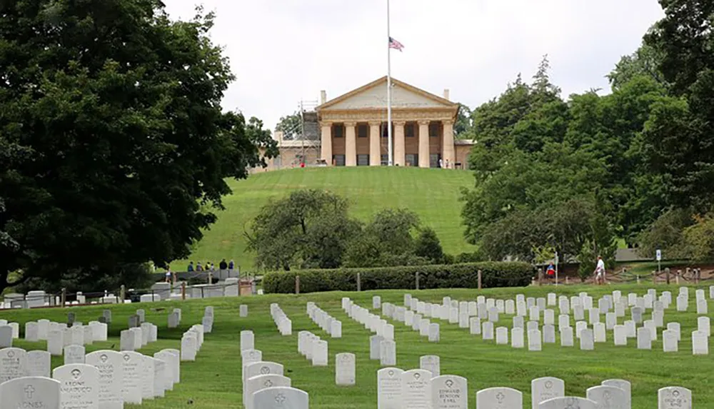 The image shows Arlington National Cemetery with rows of uniform headstones in the foreground and the Arlington House The Robert E Lee Memorial in the background under a cloudy sky