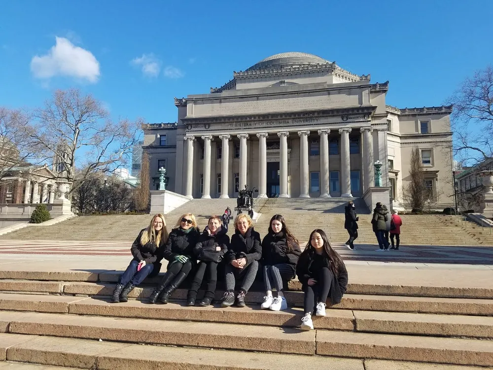 A group of people is sitting on the steps in front of the neoclassical facade of the Low Memorial Library at Columbia University on a sunny day
