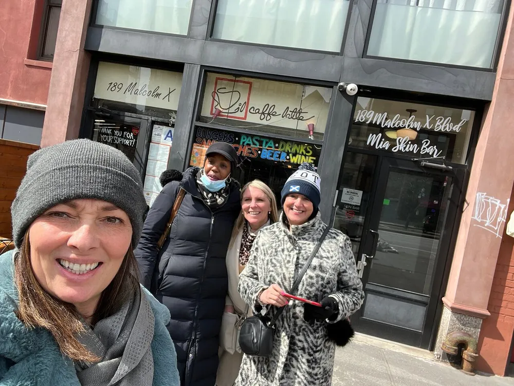 Four individuals are smiling for a selfie on a city street in front of a business establishment with signage that includes 189 Malcolm X Blvd Mia Skin Bar