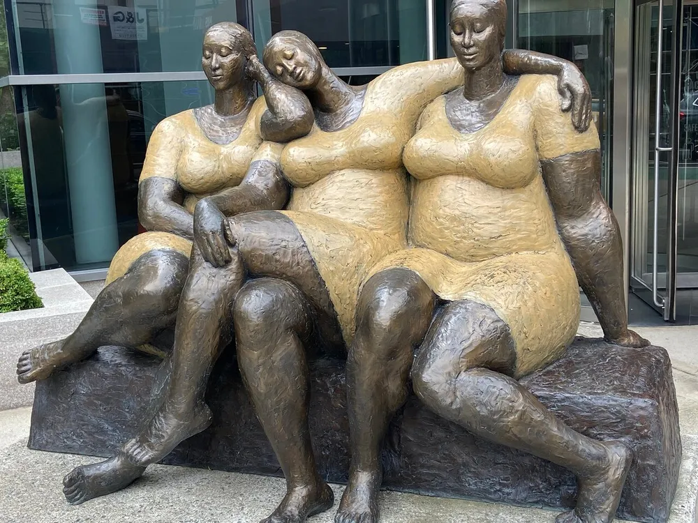 This image shows a bronze sculpture of three full-figured women sitting closely together sharing a moment of relaxation and intimacy
