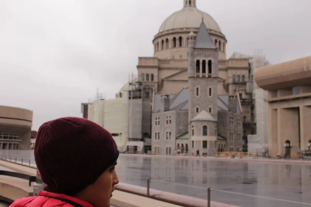 A person in a red jacket and maroon beanie is looking towards a large domed building that appears to be under renovation with a reflective surface in the foreground