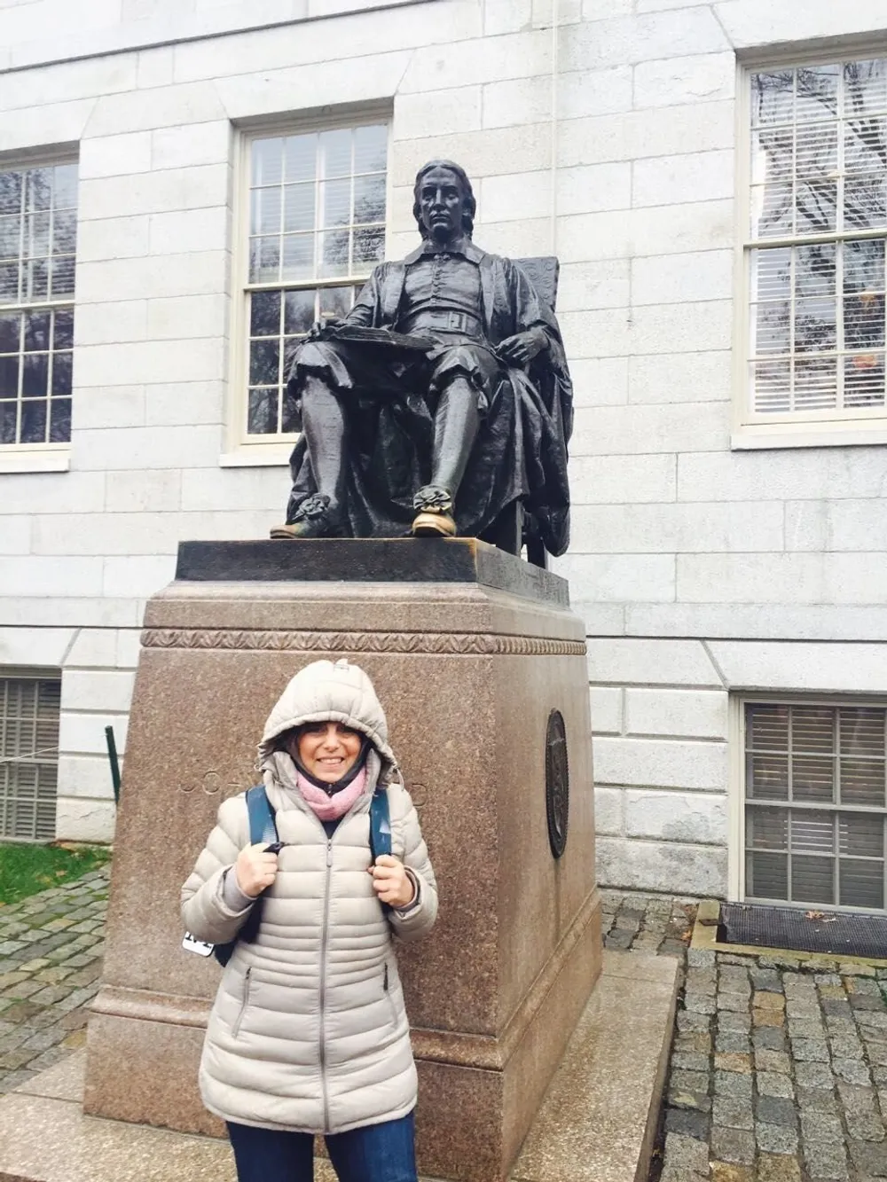 A person wearing a hooded coat and a backpack is smiling with thumbs up in front of a seated bronze statue of a historical figure on a stone pedestal