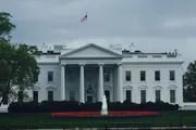 The image shows the White House, the official residence of the President of the United States, with a green lawn and an American flag flying atop the building on a cloudy day.