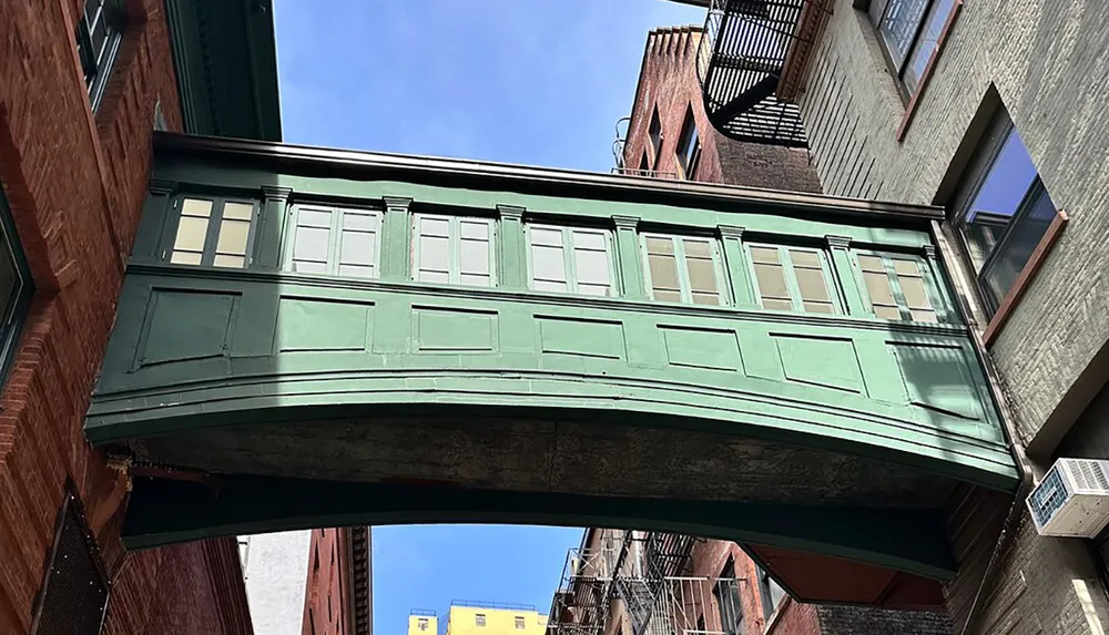 A green pedestrian bridge with windows spans between two brick buildings against a clear blue sky