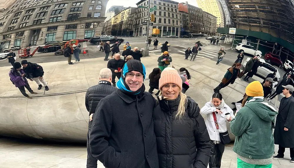 A man and a woman are smiling for a photo in front of an urban reflective sculpture with various other people walking around the area
