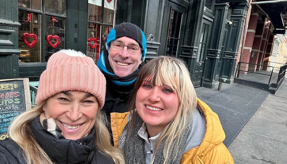 Three people are smiling for a selfie on a city street with decorations in the window behind them suggesting it might be around Valentines Day