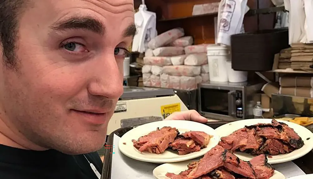 A man is smiling at the camera while showing three plates of sliced pastrami in what appears to be a deli setting