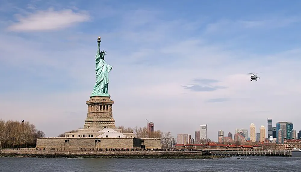 The image shows the Statue of Liberty with a helicopter flying nearby and a city skyline in the background