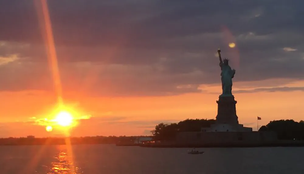 The Statue of Liberty is silhouetted against a dramatic sunset sky with a sunbeam aligning perfectly with its torch