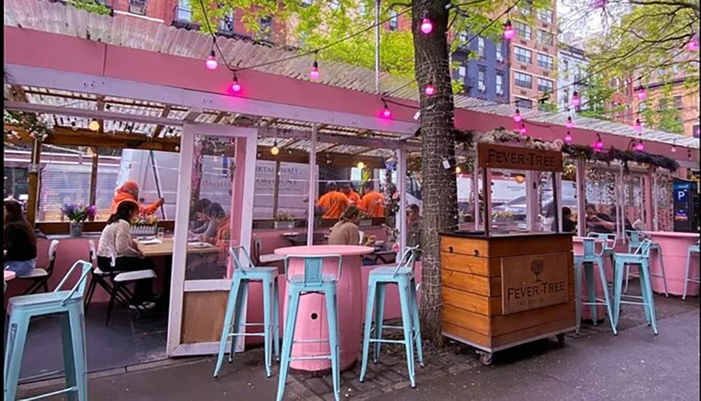 The image shows a vibrant outdoor seating area of a restaurant with pink and turquoise decor and patrons dining under a canopy adorned with hanging lights