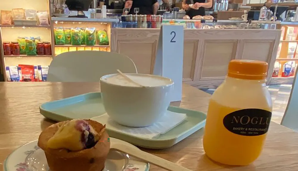 A tray with a large cup of cappuccino a muffin and a bottle of orange juice sits on a table in a cafe with a counter and products in the background