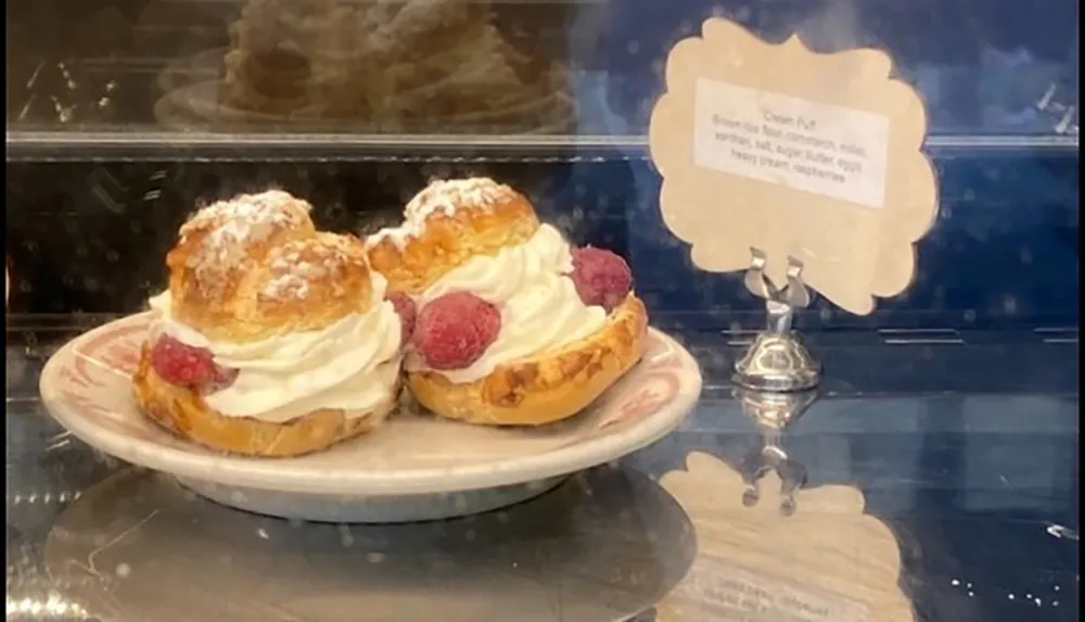 The image shows a plate with two cream puffs topped with whipped cream and raspberries displayed in a glass case next to a label listing ingredients suggesting its a bakery or dessert shop setting