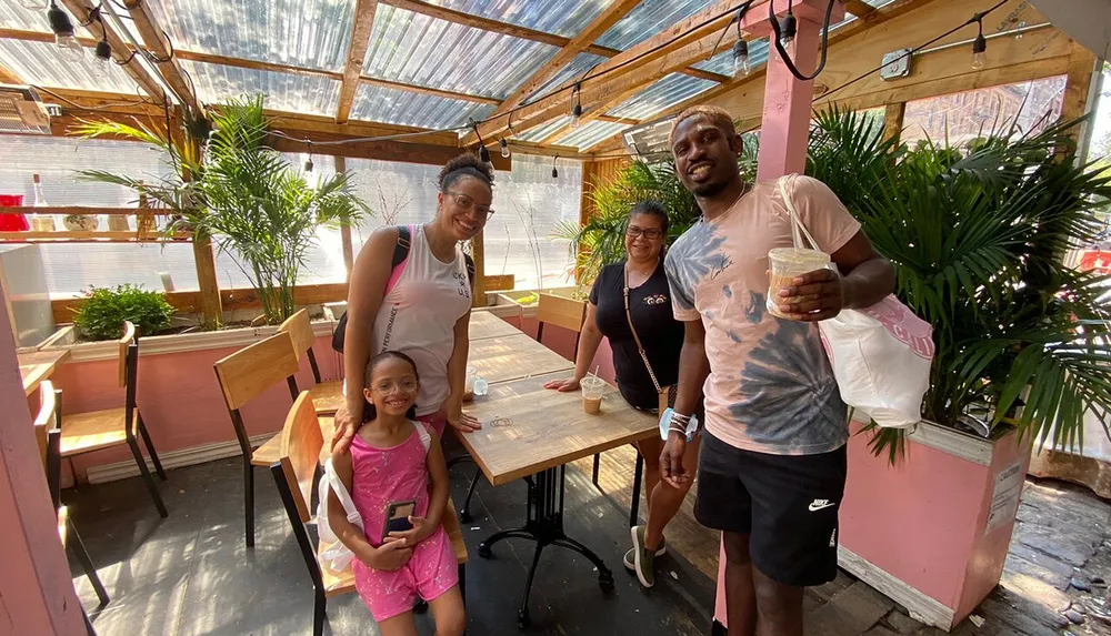 A group of four people including two adults and two children are smiling and posing together in a sunlit plant-decorated outdoor caf