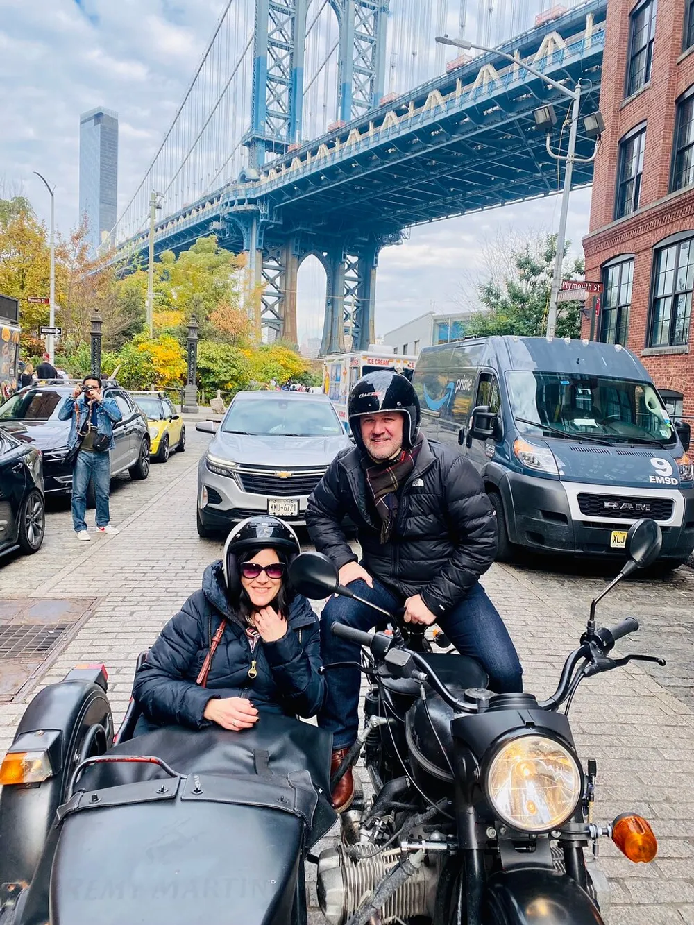 Two people are posing for a photo on a motorcycle with a sidecar in front of the Manhattan Bridge in New York City