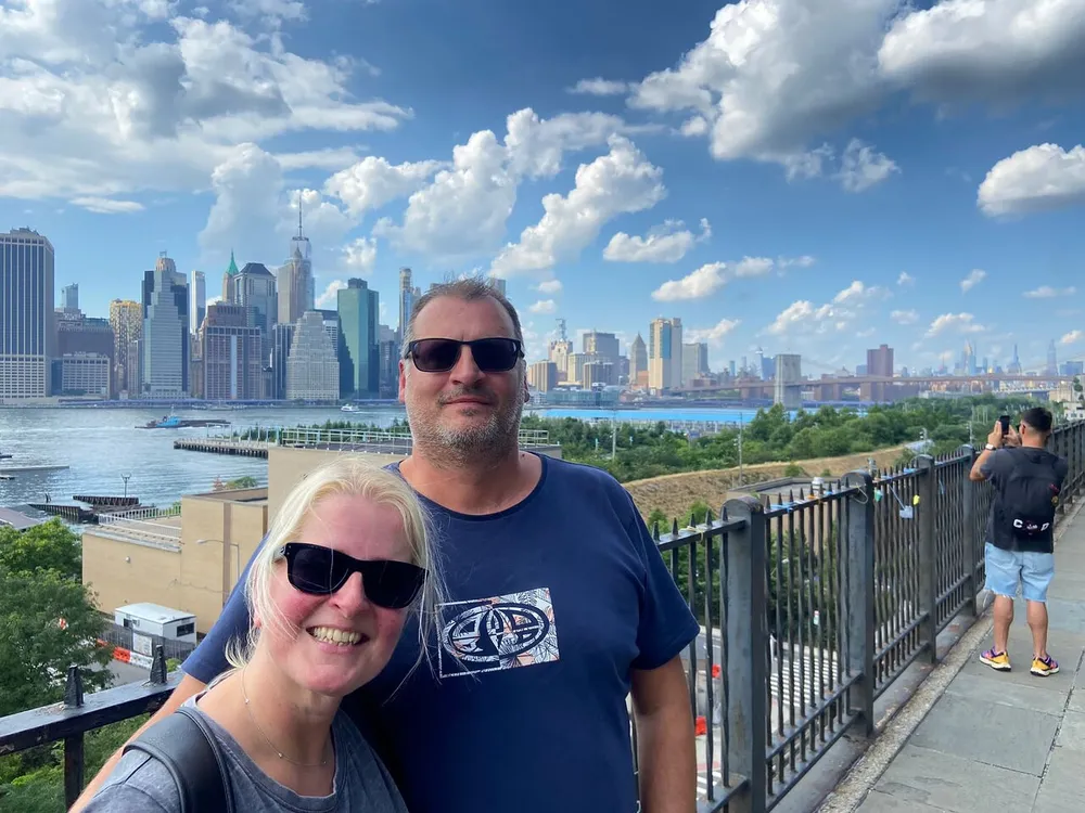 A couple is posing for a selfie with the New York City skyline in the background on a sunny day with scattered clouds