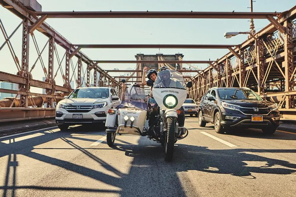A person riding a motorcycle with a sidecar is moving down a city bridge flanked by cars on one side and with complex metal structures overhead as the passenger turns to smile at the camera