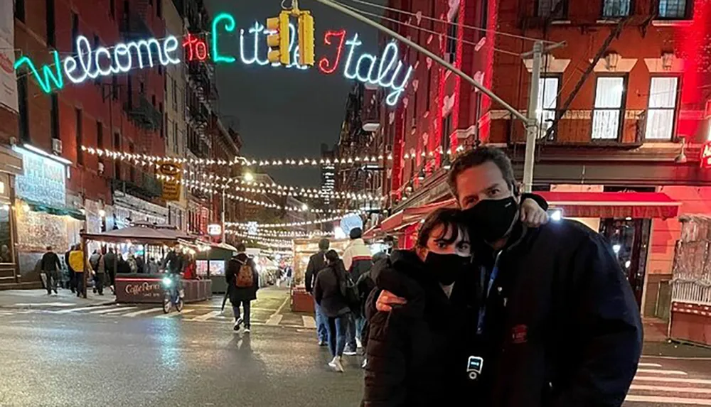 Two people wearing masks are posing for a photo in a lively street at night with a neon sign that reads Welcome to Little Italy overhead