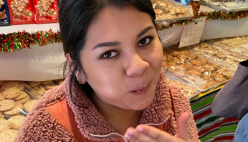 A person is making a playful expression while standing in front of a table with packaged cookies and holiday decorations