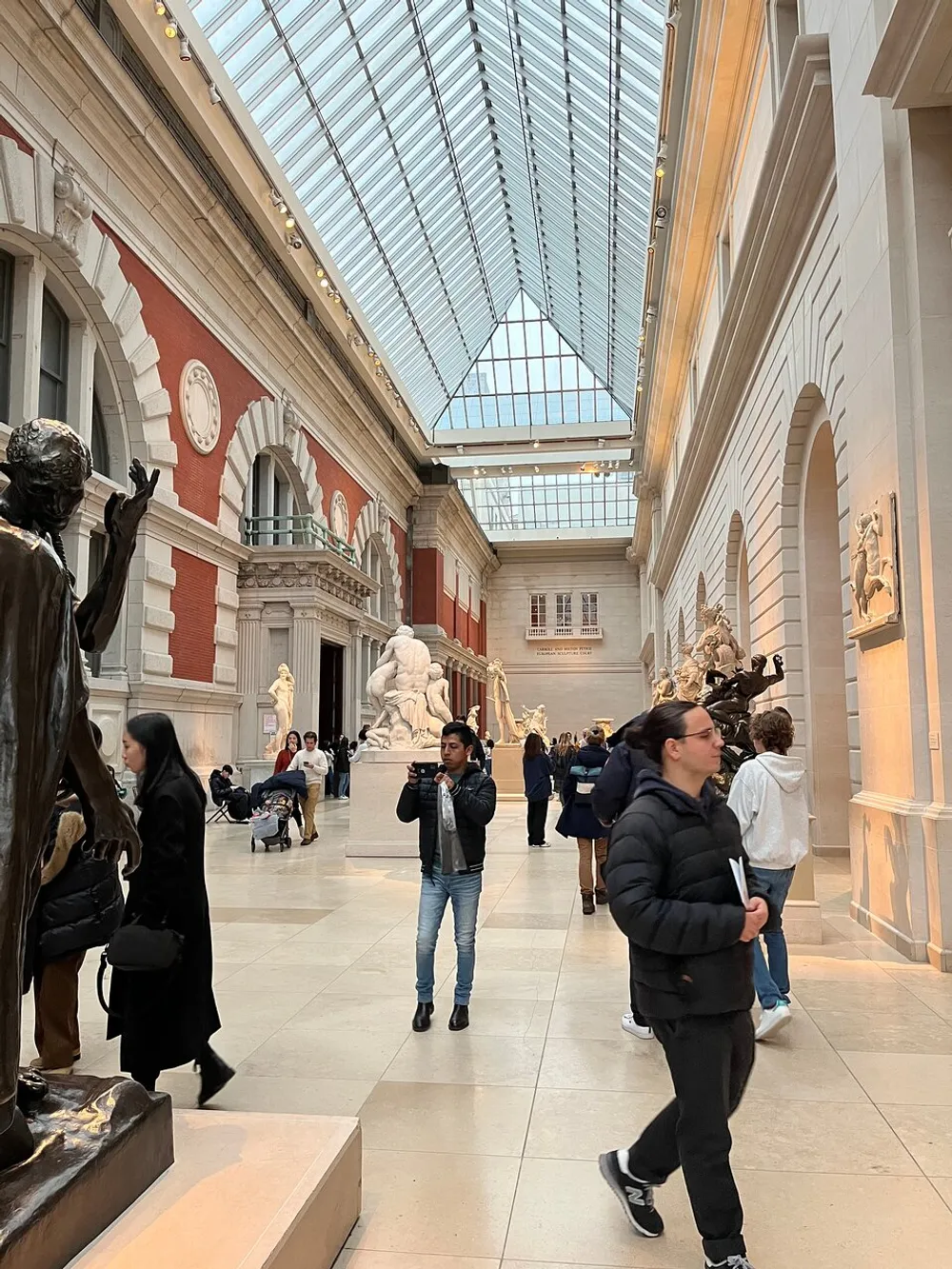 Visitors are walking through a grand hall with classical sculptures and a glass ceiling in an art museum