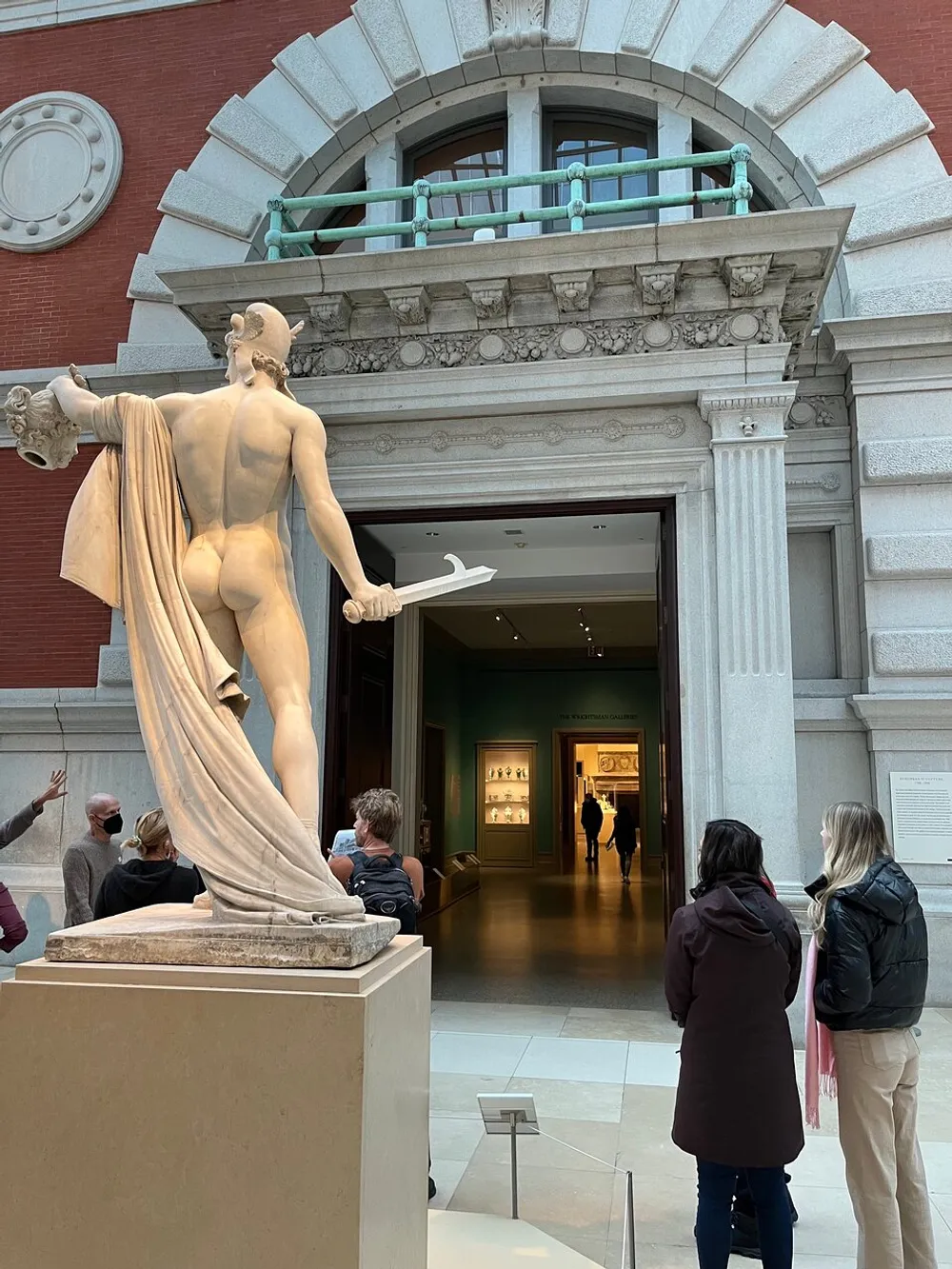 A classical statue of a nude male figure holding a sword stands prominently in a museum gallery where visitors are observing art and interacting with each other
