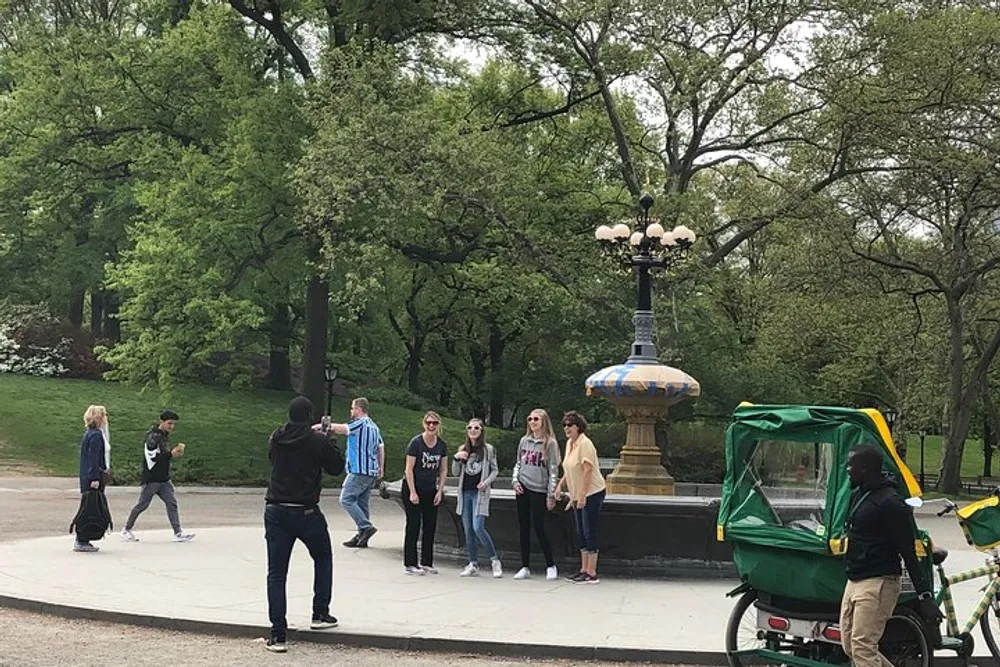 A person is taking a photo of a group next to an ornate lamp post in a park with a pedicab in the foreground and lush greenery in the background