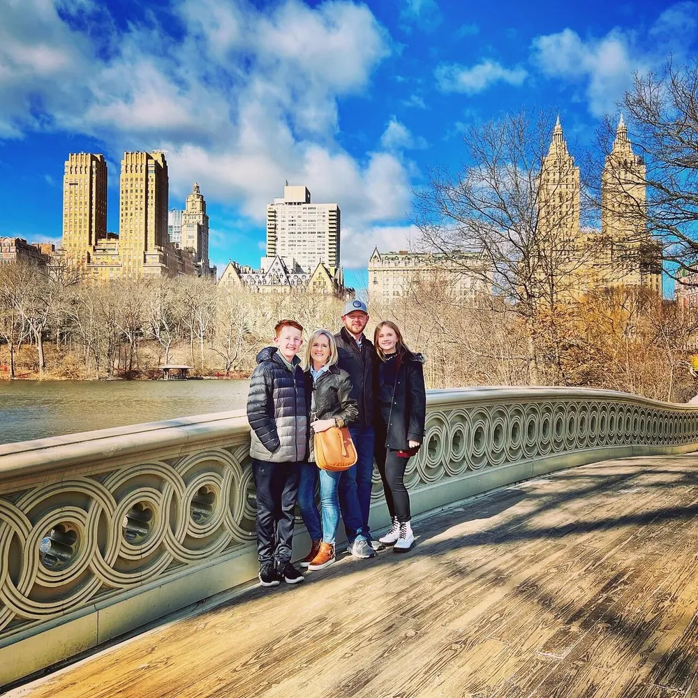 Four people are smiling for a photo on a bridge with the backdrop of a citys skyline and a park