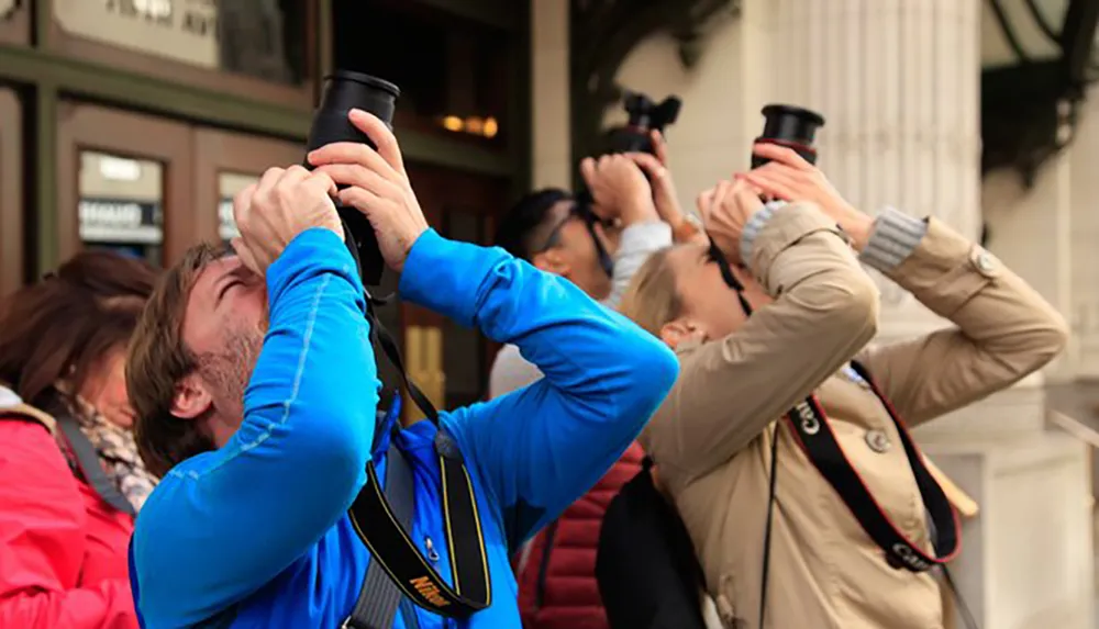 A group of people are looking up each holding binoculars or cameras to their eyes seemingly focused on something out of the frame