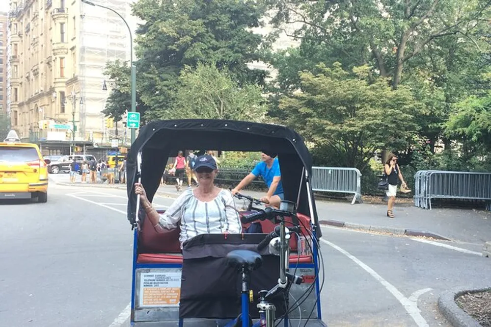 A person smiles while sitting in a pedicab on a city street next to a park
