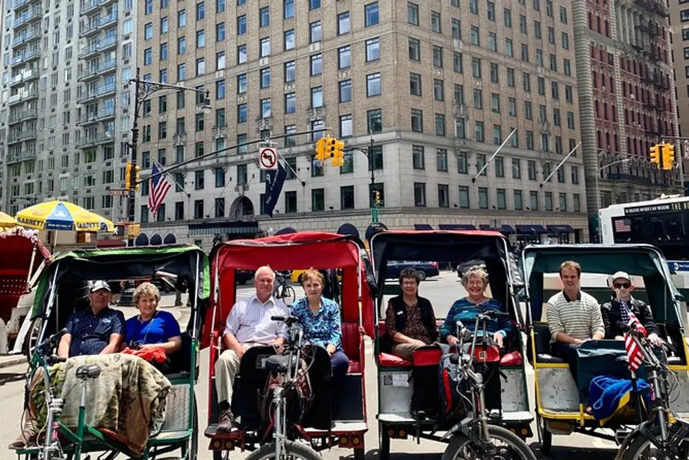 Six individuals are seated in pairs in three pedicabs lined up on a city street on a sunny day
