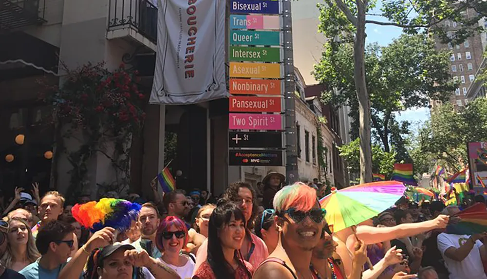 A diverse crowd celebrates at a pride event under a symbolic street sign pole with various LGBTQ community identifiers