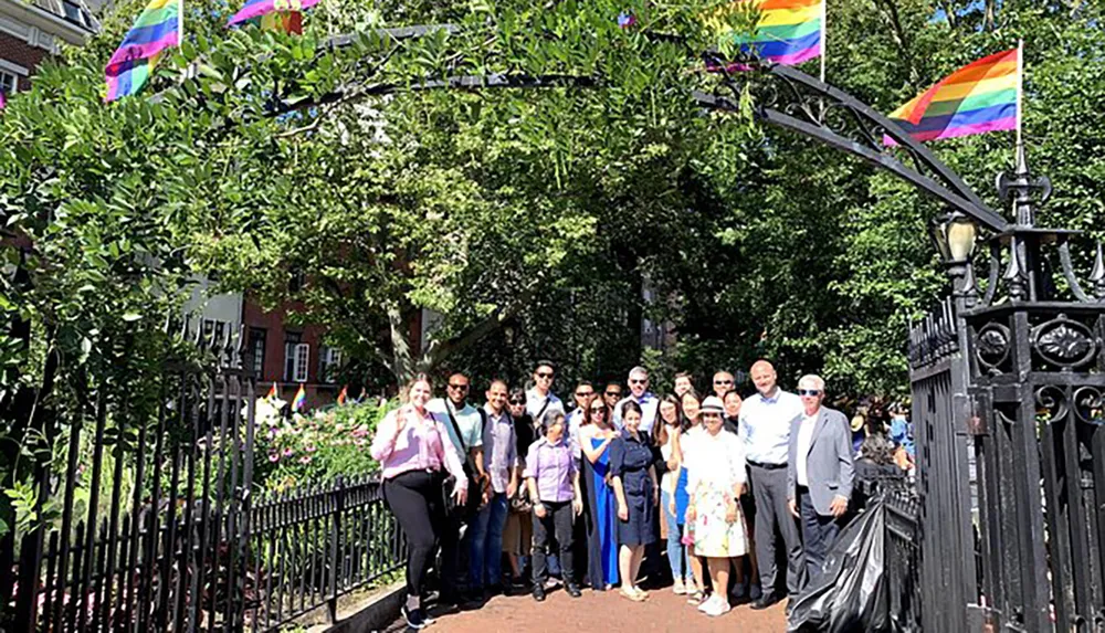 A group of individuals poses for a photo in a park-like setting with multiple rainbow pride flags in the background