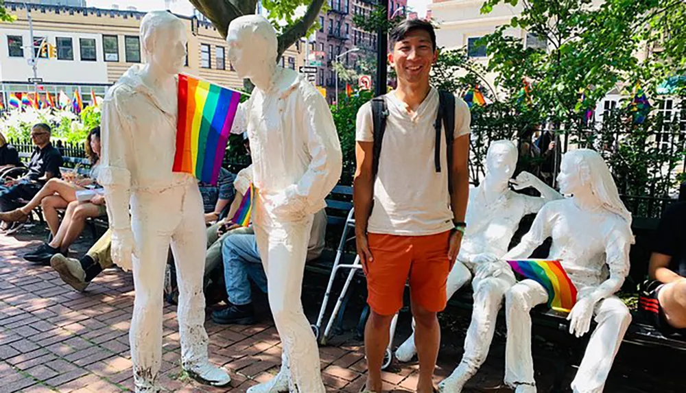 A smiling person stands beside white statues of people one of which is holding a rainbow pride flag in what appears to be a vibrant outdoor setting