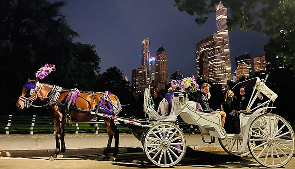 A horse-drawn carriage adorned with flowers and lights is being enjoyed by passengers on a city street at night with illuminated skyscrapers in the background