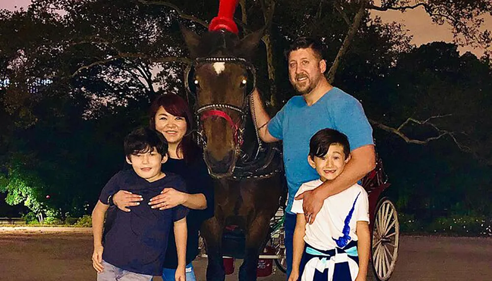 A family of four smiles for a photo alongside a brown horse decked out with a red bridle with a white carriage visible in the background in an evening outdoor setting