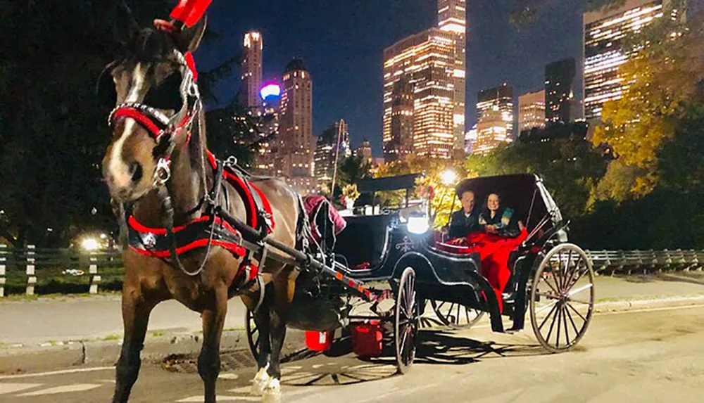 A horse-drawn carriage is seen at night on an urban path with a couple seated inside surrounded by the illuminated skyscrapers of a city backdrop