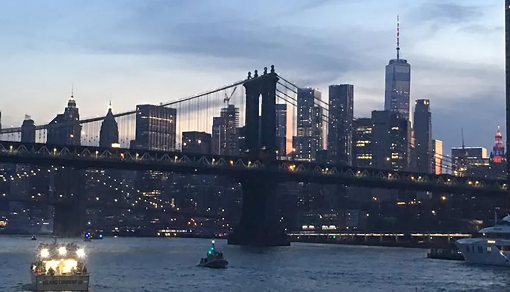 The image captures a dusk view of the Brooklyn Bridge with the New York City skyline in the background illuminated by a mix of natural and artificial lights