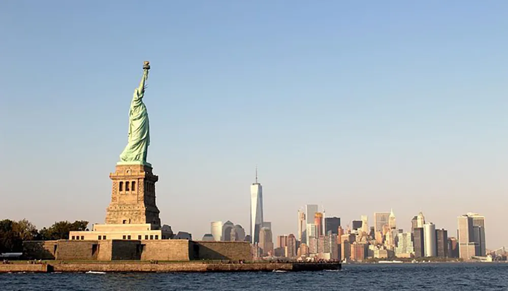The Statue of Liberty stands prominently in the foreground with the skyline of lower Manhattan including One World Trade Center in the background under a clear sky