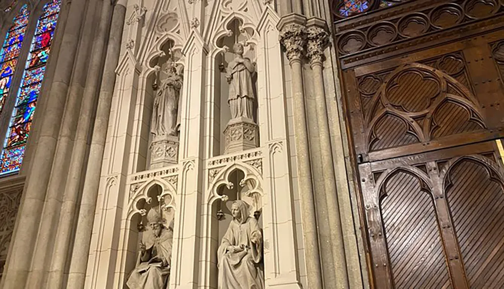 The image shows a section of a cathedrals interior featuring intricate Gothic architectural details statues set in niches and part of a stained glass window